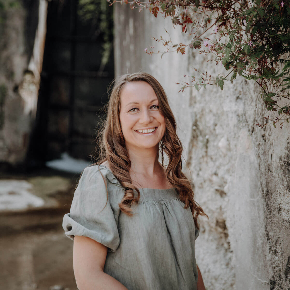 Frau mit langen, braunen Haaren lächelt, trägt ein graues Kleid und steht neben einer alten Steinmauer mit Pflanzen.