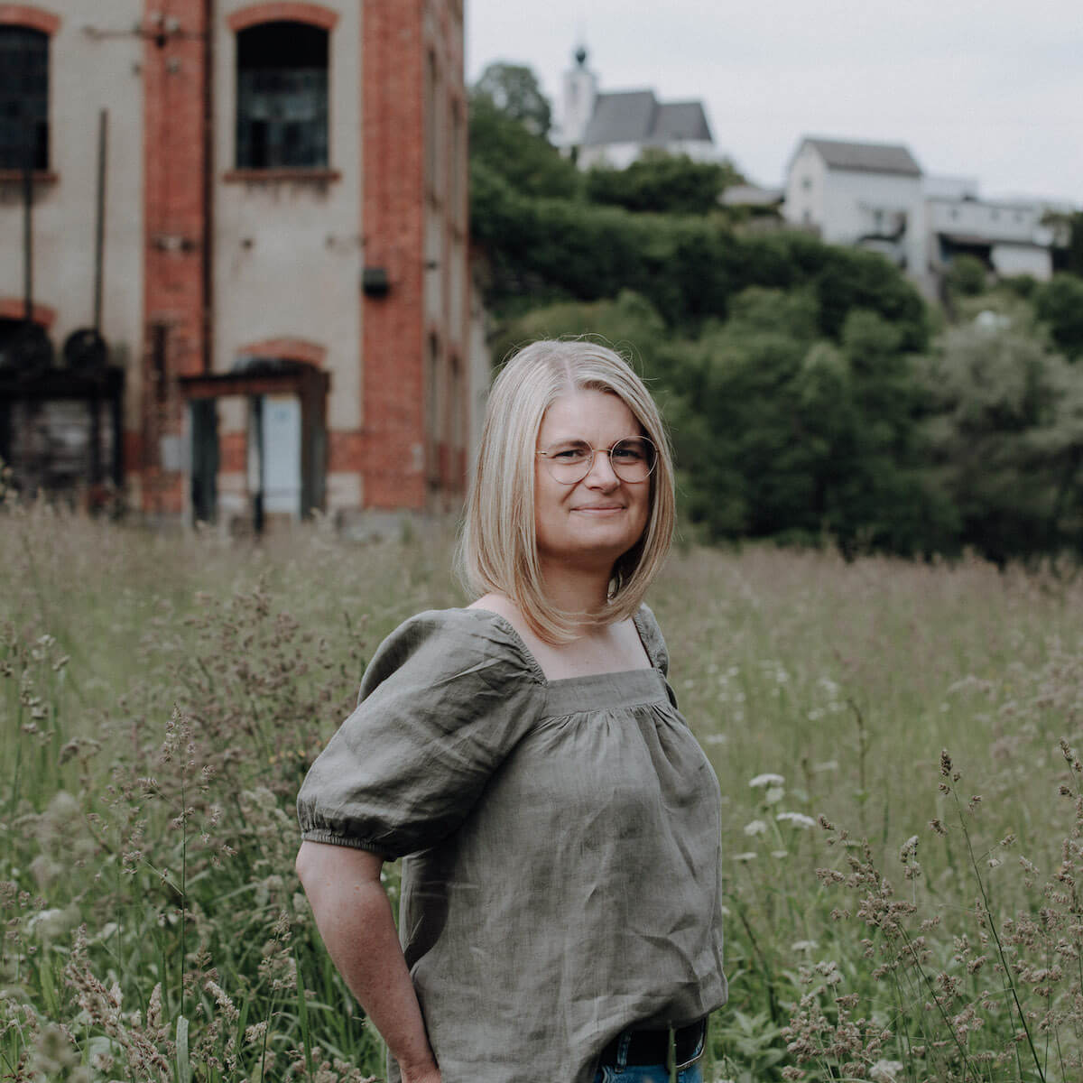 Eine Frau mit Brille steht in einem Feld vor einem alten Backsteingebäude, umgeben von Bäumen und Häusern im Hintergrund.