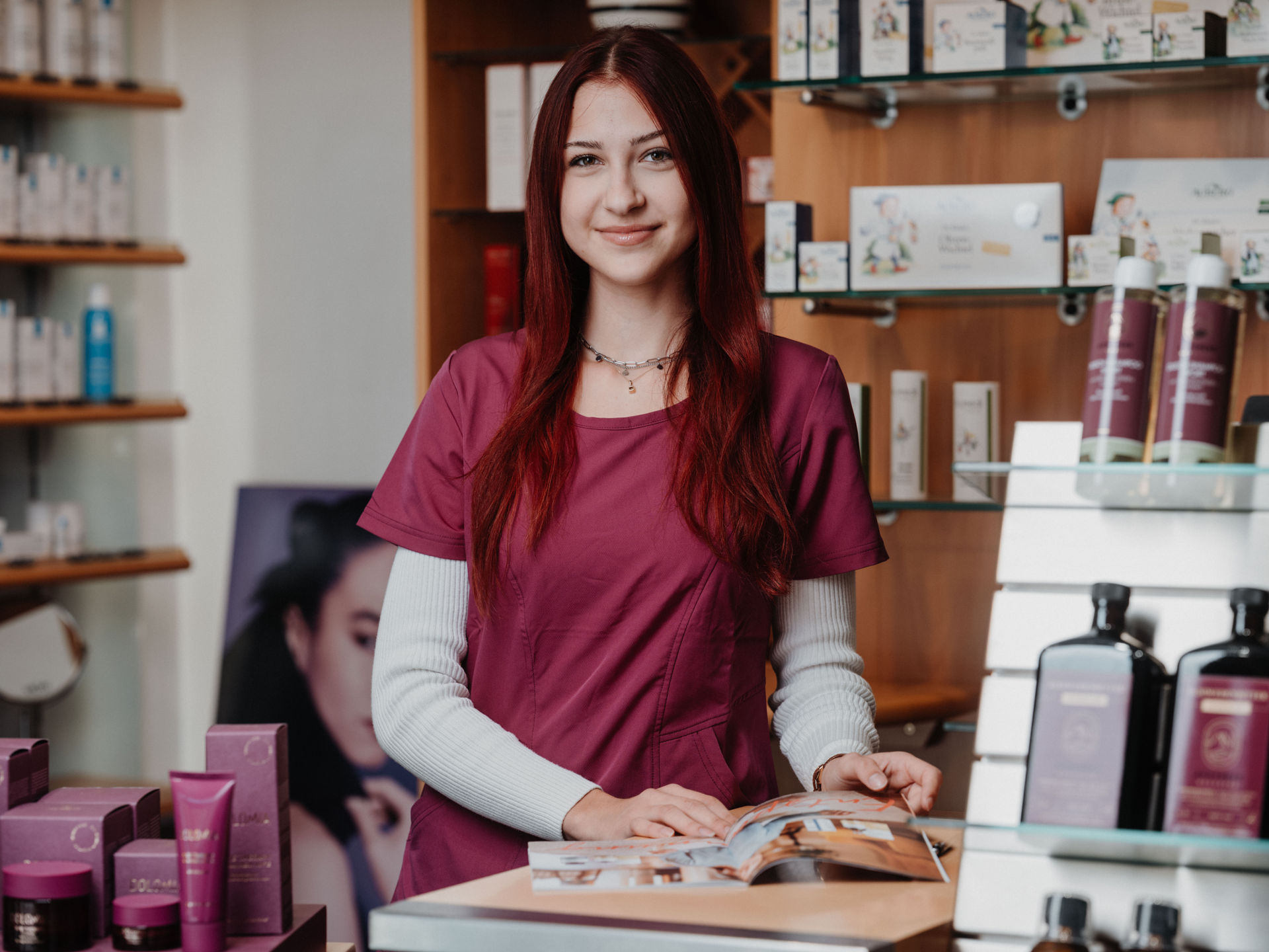 Eine Frau mit roten Haaren steht lächelnd in einem Geschäft für Hautpflegeprodukte, vor einem Regal mit verschiedenen Artikeln.