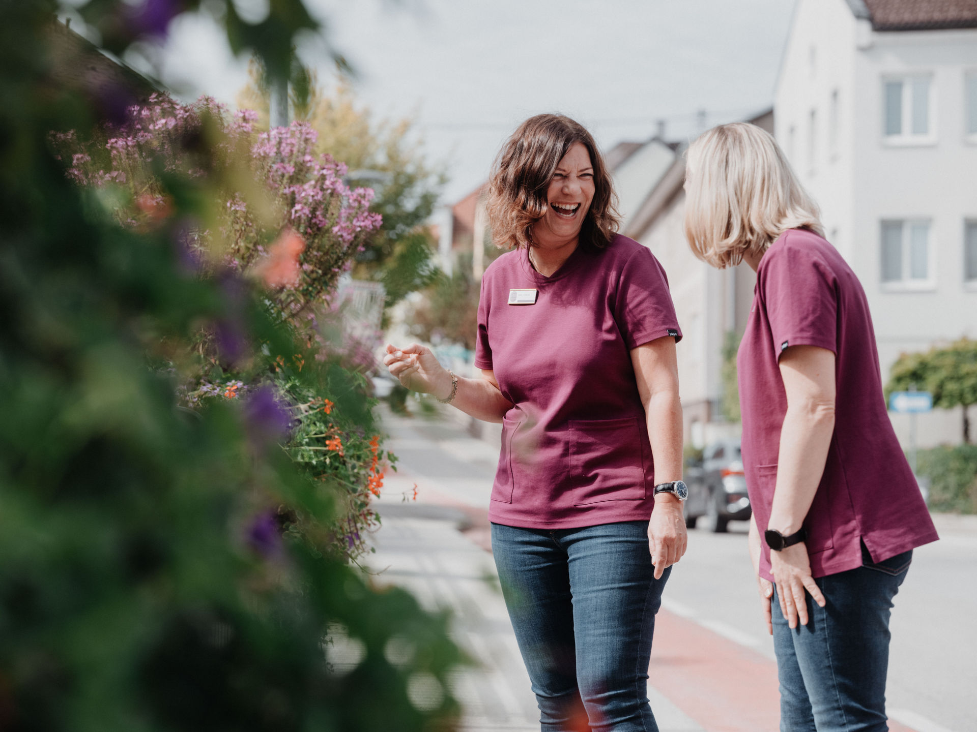 Zwei lachende Frauen in lila Oberteilen stehen auf einer Straße und schauen auf Blumen am Straßenrand.