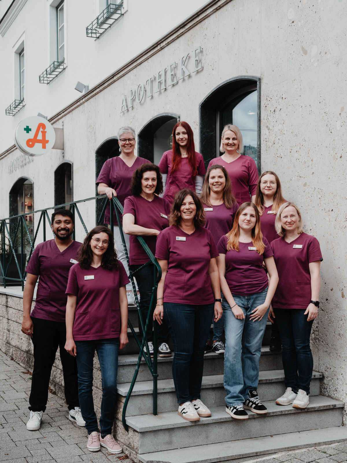 Eine Gruppe von zehn Personen in lila Oberteilen steht vor einer Apotheke auf einer Treppe und lächelt in die Kamera.