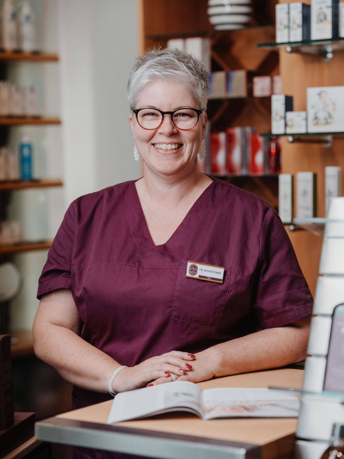 Eine lächelnde Frau mit Kurzhaarfrisur und Brille trägt eine lila Uniform und steht in einem Geschäft mit Produkten im Hintergrund.