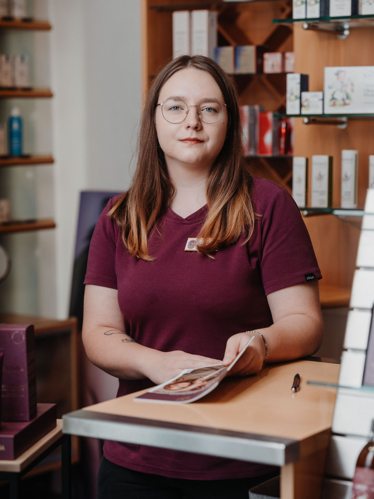 Eine Frau mit Brille und lila T-Shirt steht an einem Tresen, hält eine Broschüre, im Hintergrund sind Regale mit Produkten.