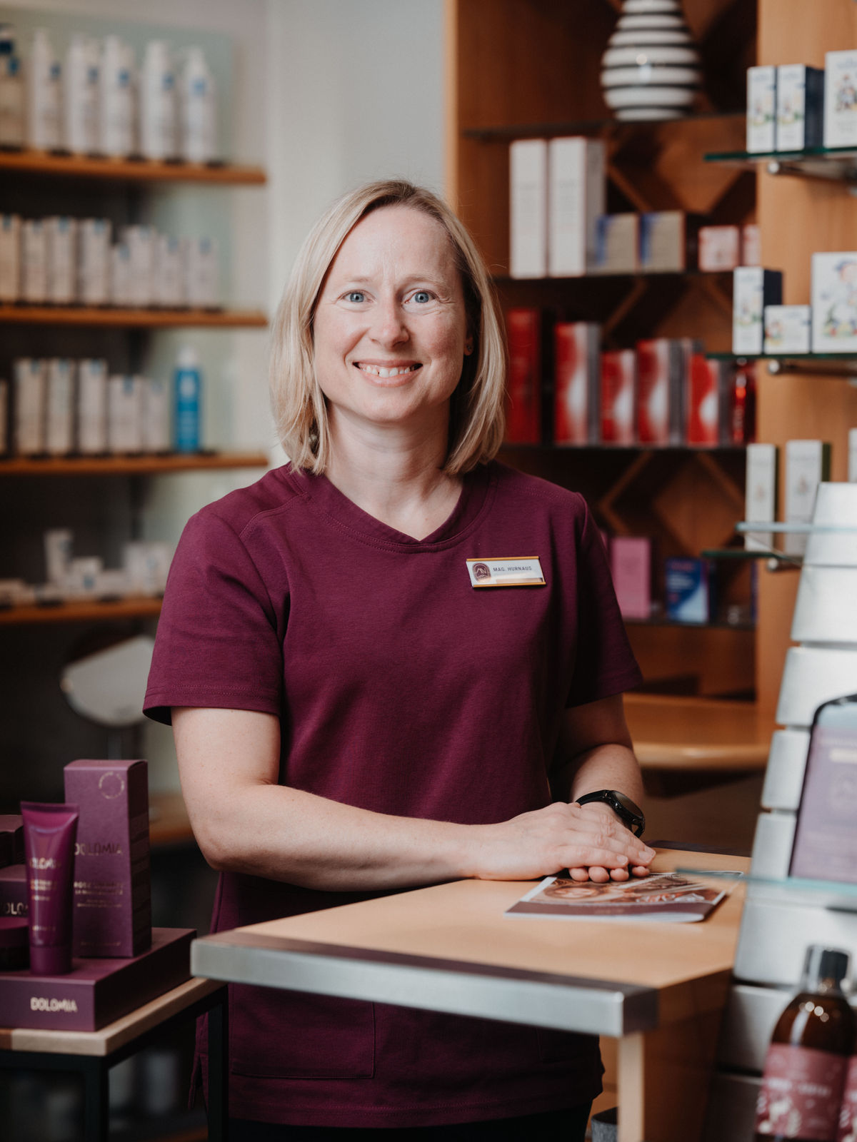 Eine lächelnde Frau in bordeauxfarbenem Oberteil steht an einem Tresen in einem Geschäft mit Kosmetikprodukten im Hintergrund.