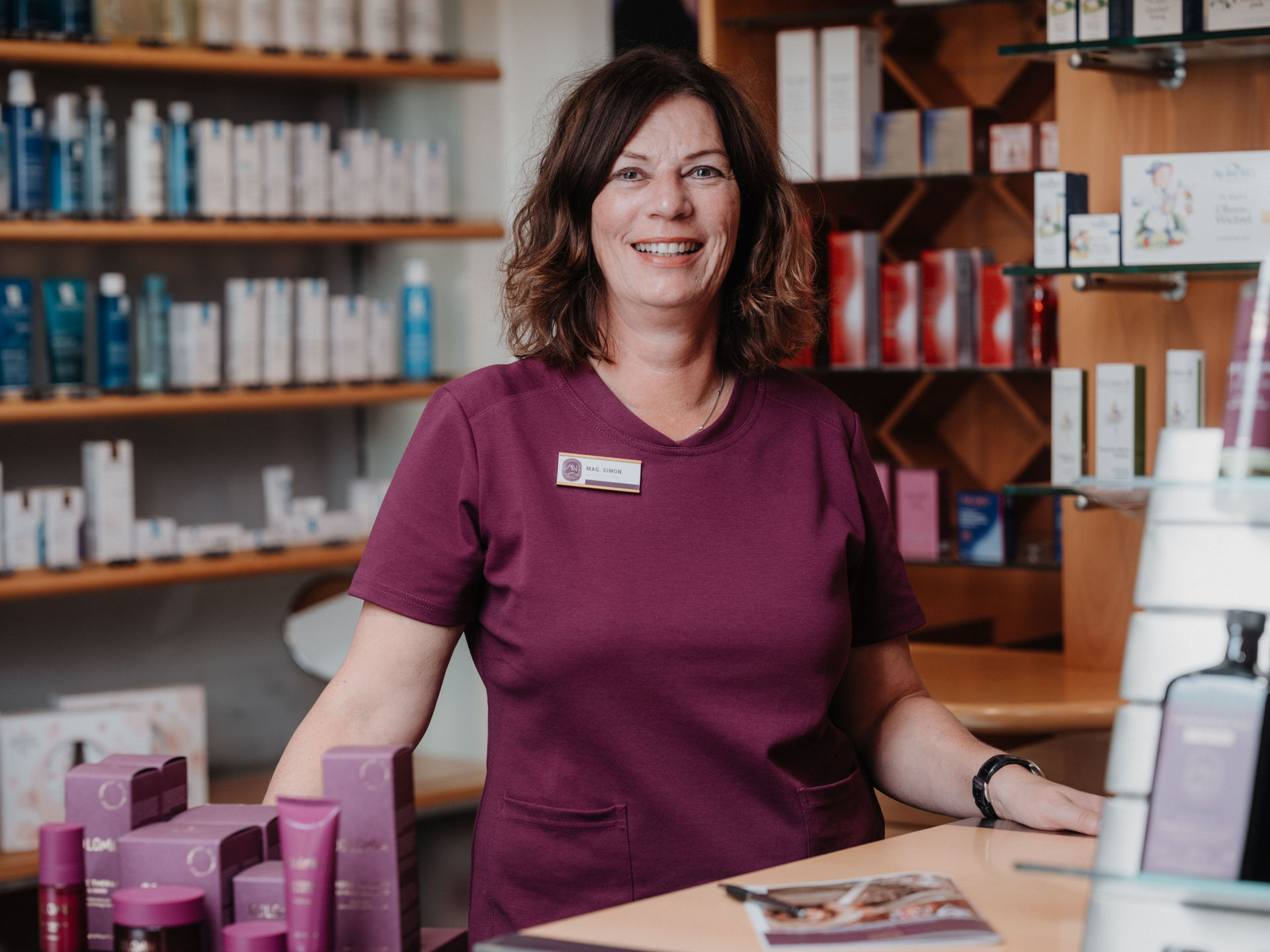 Eine Frau in lila Kleidung steht lächelnd hinter einem Tresen, neben einem Blumenstrauß, in einem Geschäft mit Gesundheitsprodukten.