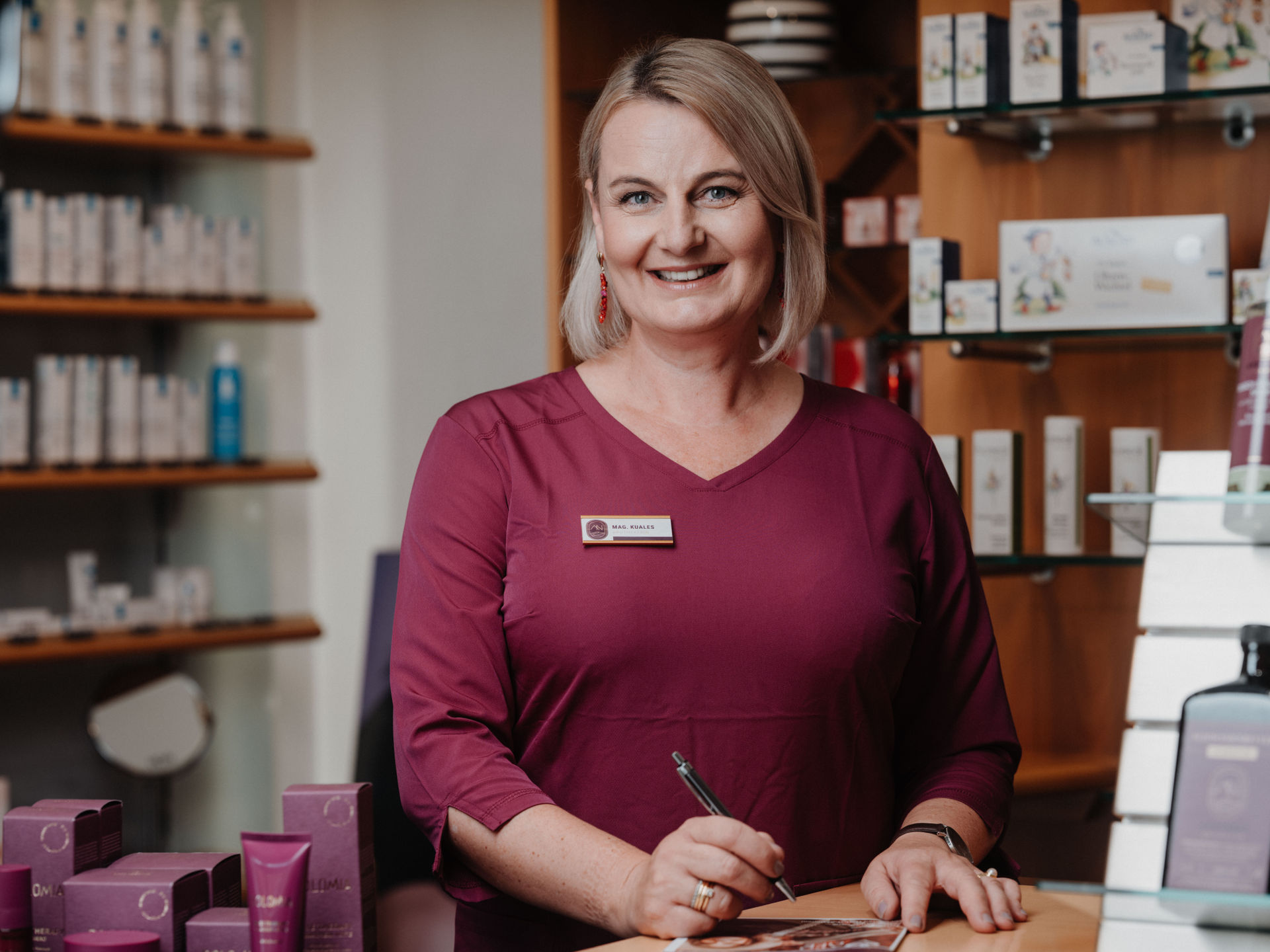 Eine Frau in einer lila Bluse steht lächelnd an einem Verkaufstresen, umgeben von Kosmetikprodukten in einem Geschäft.