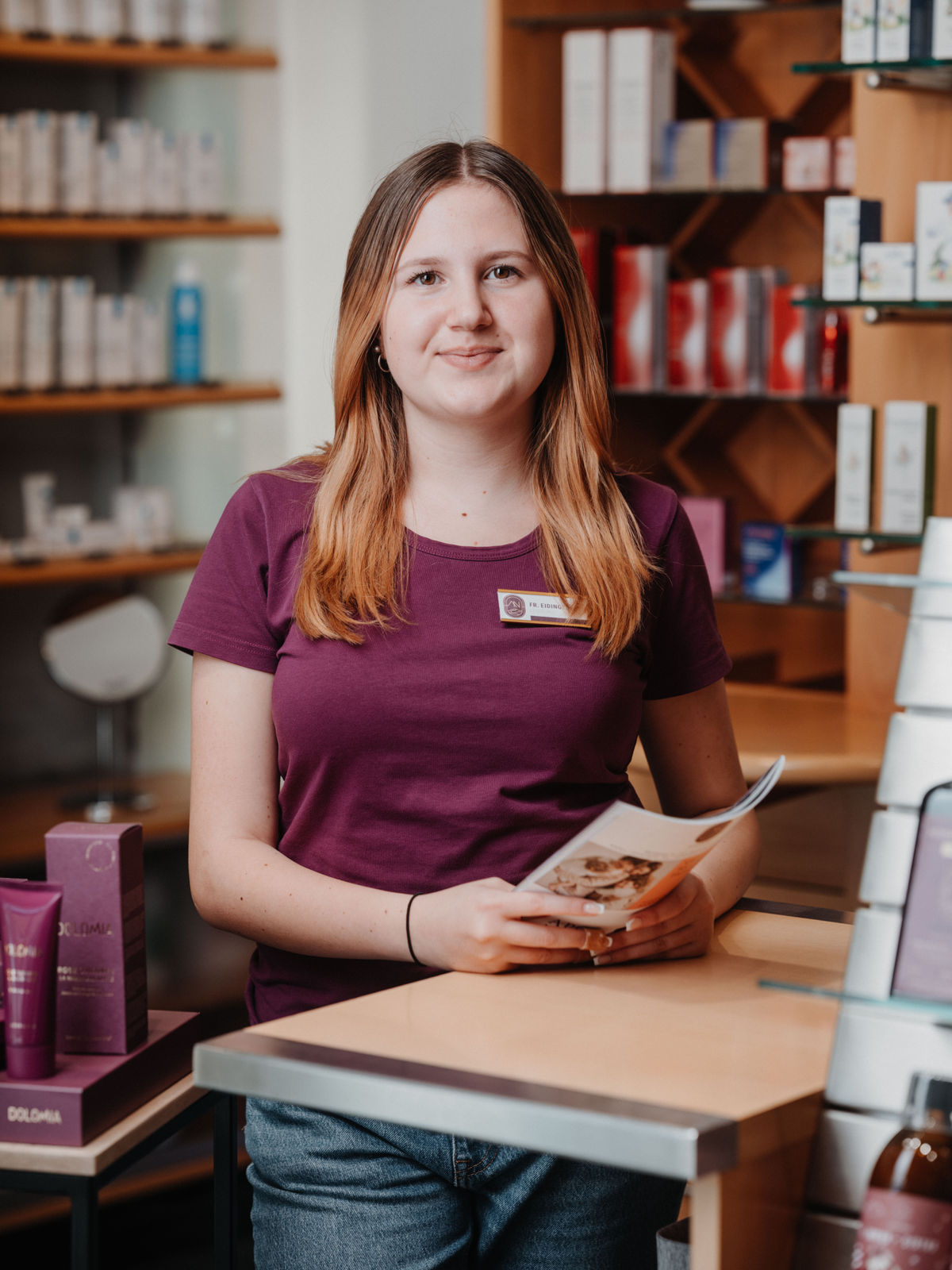 Eine junge Frau in lila T-Shirt steht in einer Apotheke, hält eine Broschüre und lächelt. Im Hintergrund sind Regale mit Produkten.