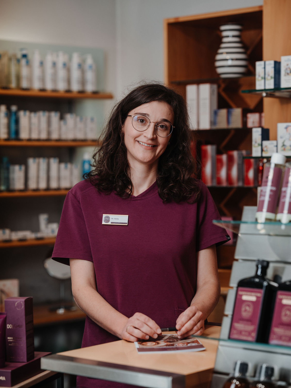Eine Frau mit Brille und Namensschild steht lächelnd in einem Geschäft mit Kosmetikprodukten und einem Regal im Hintergrund.