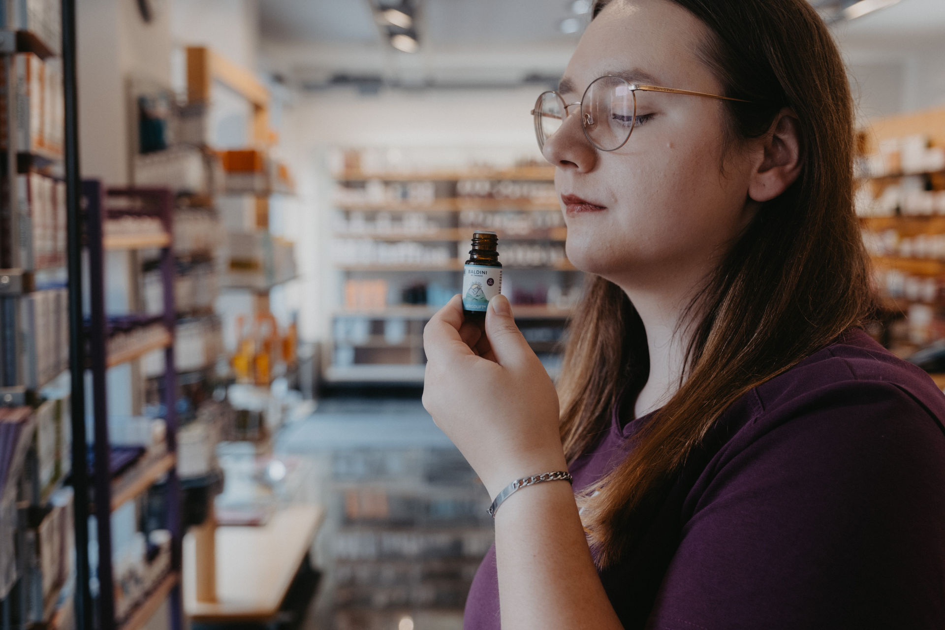 Eine Frau mit Brille riecht an einem Fläschchen mit ätherischem Öl in einem Geschäft mit Regalen voller Produkte im Hintergrund.