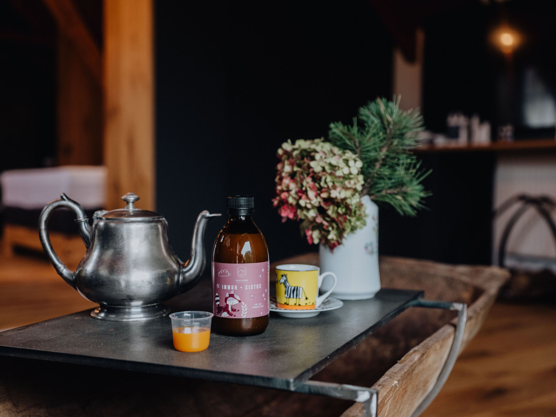 Eine silberne Teekanne, eine Flasche mit der Aufschrift „Immun + Cistus“, eine kleine Tasse und ein Blumenstrauß auf einem Tisch.
